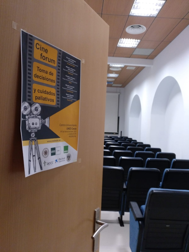 CINEFORUM "Toma de decisiones en Cuidados Paliativos" Tablón con un cartel y bucacas a la derecha